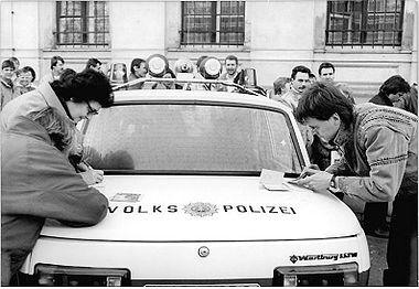 Ein Auto der Volkspolizei dient diesen jungen Leipzigern als Schreibunterlage zum Ausfüllen der Reiseformulare. - Foto: Kluge, Wolfgang - Deutsches Bundesarchiv